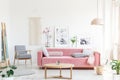 Real photo of a pink couch with pillows standing behind a table, next to an armchair, in front of a shelf with posters and flower