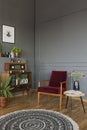 Real photo of a red armchair standing between a stool with flowers and shelf with books, radio and plants in retro living room in Royalty Free Stock Photo