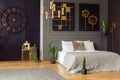 Real photo of a spacious bedroom interior with grey walls, clock, paintings, plants, bed and golden accents