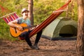 Handsome young man in hat play guitar while lying in hammock , camping on background