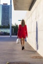 Rear image of a blonde woman wearing red jacket white handbag while walking in the city