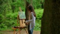 Rear middle view of a young artist woman painting a landscape in a summer forest