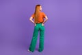 Rear photo of red hair woman in orange t shirt put arms into trousers looking empty space with smile isolated on violet