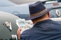 Rear shot of a caucasian man in a hat sat beside classic cars reading adverts for classic cars in a newspaper. Over shoulder shot