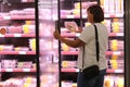 Rear view of adult female customer opens big glass door in supermarket's fridge and chooses packaged fresh chilled