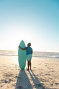 Rear view of african american senior woman with surfboard looking at sea under blue sky at sunset Royalty Free Stock Photo