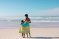 Rear view of african american young mother and daughter with towel standing at beach against sky Royalty Free Stock Photo