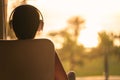 Rear view of alone sad woman in headphones listening music sitting on chair near window and looking on palm trees sea beach at sun Royalty Free Stock Photo