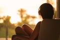 Rear view of alone sad woman in headphones listening music sitting on chair near window and looking on palm trees sea beach at sun Royalty Free Stock Photo