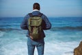 Rear view of a backpacked male tourist looking at waves splashing and crushing on the headland, standing on the cliff Royalty Free Stock Photo