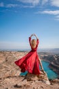 Rear view of a beautiful sensual woman in a red long dress posing on a rock high above the lake in the afternoon Royalty Free Stock Photo