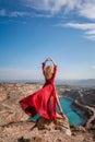 Rear view of a beautiful sensual woman in a red long dress posing on a rock high above the lake in the afternoon Royalty Free Stock Photo