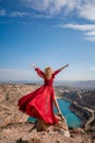 Rear view of a beautiful sensual woman in a red long dress posing on a rock high above the lake in the afternoon Royalty Free Stock Photo