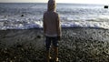 Rear view of a boy in striped jacket standing on a pebble ocean shore. Creative. Child looking at waves on a sunny day. Royalty Free Stock Photo