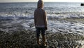 Rear view of a boy in striped jacket standing on a pebble ocean shore. Creative. Child looking at waves on a sunny day. Royalty Free Stock Photo