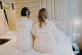 Rear view of bride and her maid of honor sitting on the bed with labeled clothes, before wedding