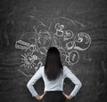 Rear view of brunette who is looking at the black chalkboard with drawn sketch of business icons, exclamation and question marks. Royalty Free Stock Photo