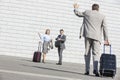 Rear view of businessman carrying luggage waving hand to colleagues