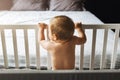 Rear view. A child alone is standing at home in a crib, holding on to its side and looking at an empty bed in the background