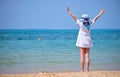 Rear view of child girl in white dress and hat standing barefoot with arms raised up on sandy beach enjoying tropical Royalty Free Stock Photo