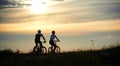 Rear view of couple cyclists riding along the road among grass with wildflowers under evening sky