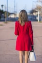 Rear view of an elegant woman wearing red jacket, skirt and holding a white handbag while walking in the street in a sunny day Royalty Free Stock Photo
