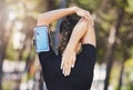 Rear view of fit young female athlete stretching her hands behind her back while listening to music on smart phone in Royalty Free Stock Photo