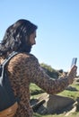Rear view of a good looking long haired young man taking selfie or having video call with his smartphone in the mountain
