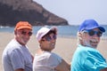 Rear view of group of three friends sitting on the beach, retired adult people in outdoor vacation at sea smiling enjoying sun and Royalty Free Stock Photo