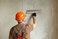 Rear view of handyman worker in overalls and protective helmet plastering concrete wall with putty using a big putty Royalty Free Stock Photo