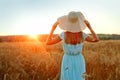 A rear view of a happy young woman in a blue summer dress and a straw hat, standing on a yellow farmer`s meadow with ripe golden Royalty Free Stock Photo