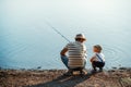 A rear view of mature father with a small toddler son outdoors fishing by a lake.