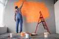 Rear view of painter man painting the wall in orange color with paint roller, empty space with red ladder. Royalty Free Stock Photo