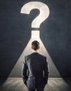 Rear view puzzled businessman in a dark room in front of a wall with illuminated interrogation mark. Finding answer to his Royalty Free Stock Photo