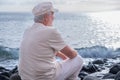 Rear view of senior caucasian man sitting on the pebble beach relaxing and looking at horizon over water. Elderly white haired man Royalty Free Stock Photo