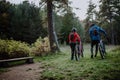 Rear view of senior couple bikers walking and pushing e-bikes outdoors in forest in autumn day.