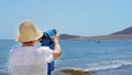 Rear view of senior woman traveler wearing hat and holding blue telescope binocular adjusting lens Royalty Free Stock Photo