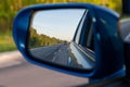 Rear view in the side mirror of a blue sedan with the reflection of a gray truck on an asphalt road on a summer day with green