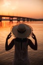 rear view of the silhouette of a girl holding the brim of a straw hat by the river and an urban landscape at an orange Royalty Free Stock Photo