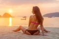 Rear view of slim female model sitting on seashore wearing bikini looking away during sunrise with sun path reflected Royalty Free Stock Photo