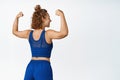 Rear view of sports woman shows strong arms, flexing biceps in sportsbra and shorts, standing against white background