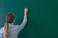 Rear view of a student or teacher with long brunette hair writing on a blank green blackboard or chalkboard with copyspace Royalty Free Stock Photo