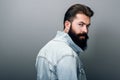 Rear view of stylish brutal young Caucasian male with thick beard wearing trendy denim jacket, staring at camera with serious look