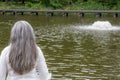 Rear view of a thoughtful pensive mature woman with long grayish straight hair, wearing a white blouse looking at a fountain in a