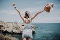 Rear view of tourist woman on cliff rock by blue sea and sky, stretching arms up, outdoors nature freedom. Healthy female enjoying