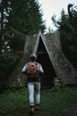 Rear view traveler man with backpack coming to old vintage wooden hut in dark woods Royalty Free Stock Photo