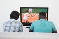 Rear view of two friends sitting at home together watching tennis on tv