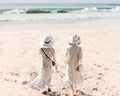 Rear view of two young women in long dresses and hats walking along sandy beach