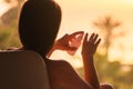 Rear view of woman applying cream sitting on armchair near panoramic window and looking on palm trees sea beach at sunset. Royalty Free Stock Photo