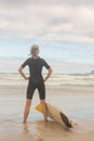 Rear view of woman with hand on hip standing by surfboard on shore Royalty Free Stock Photo